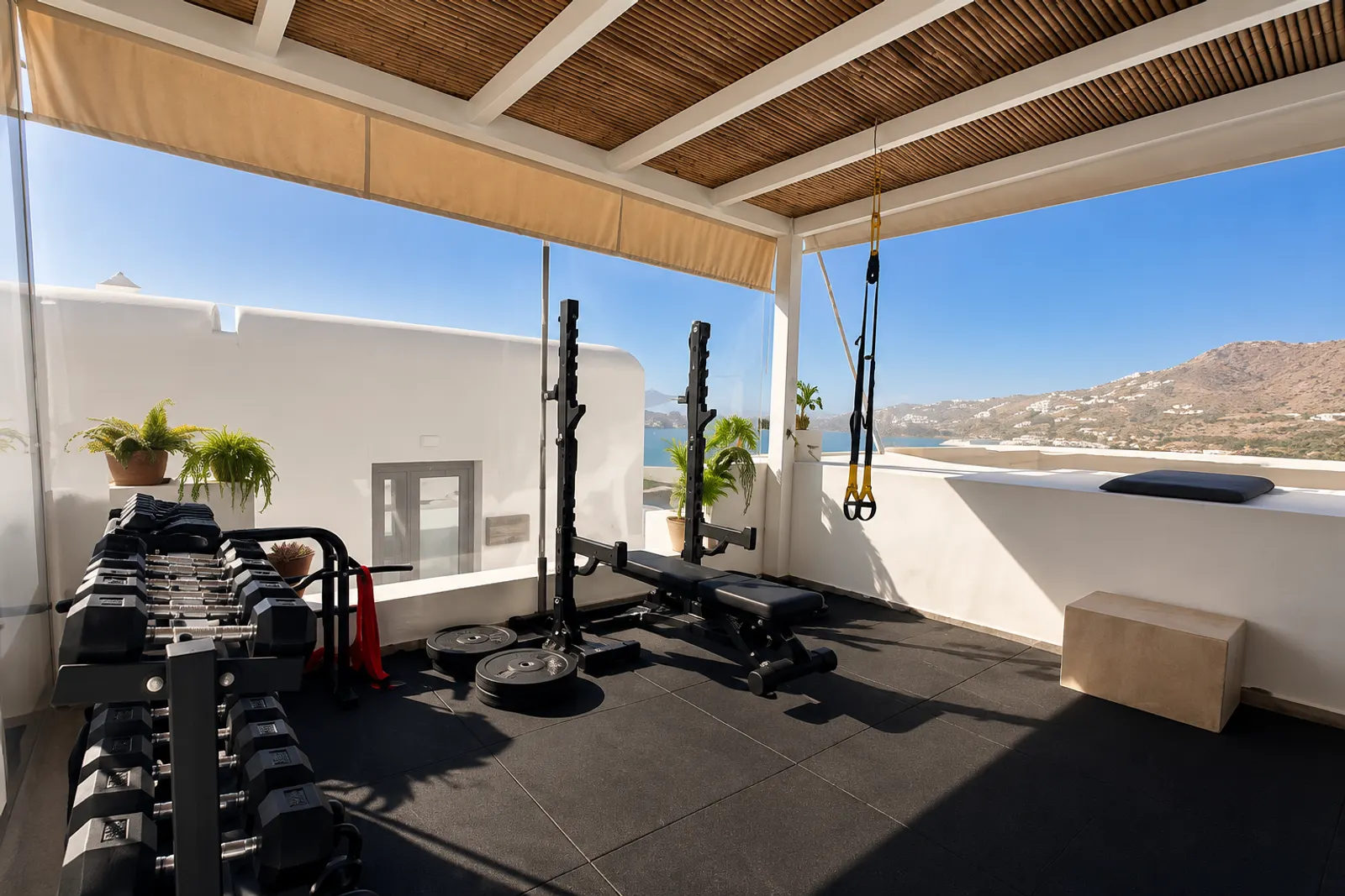 Open-air rooftop gym with dumbbells and a training bench overlooking the sea and hills of Mykonos